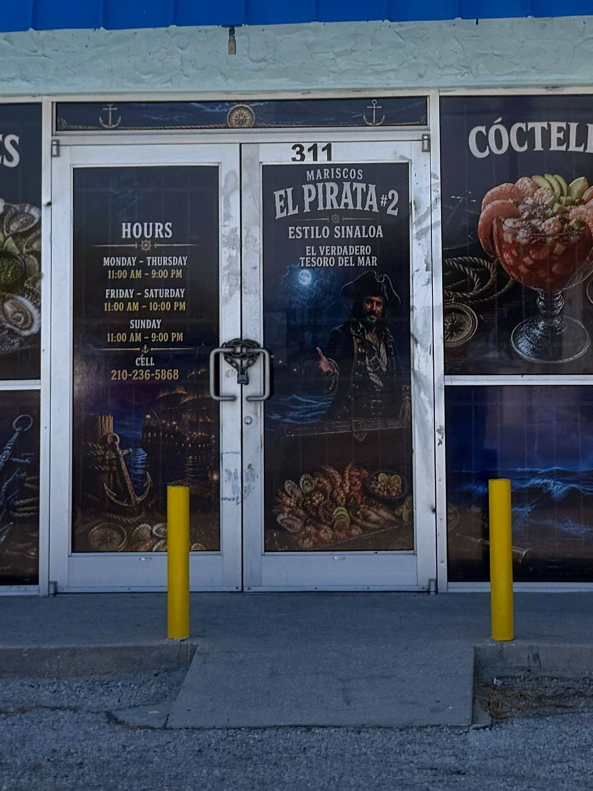 La puerta de Mariscos El Pirata #2: el pirata pintado sobre el mar de noche, señalando hacia adentro, con el horario y el número 311 al lado